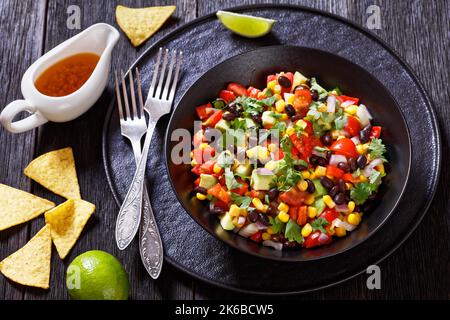 Cowboy-Kaviar, texas-Kaviar mit schwarzer Bohne, Tomaten, Avocado, roter Paprika, Mais, Koriander in schwarzer Schale auf dunklem Holztisch mit Nachos und d Stockfoto
