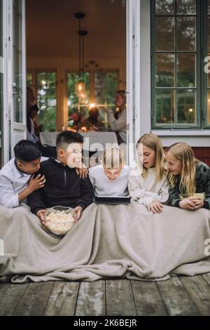 Aufgeregt Freunde mit Decke und Popcorn beobachten Tablet-PC, während sie auf der Veranda sitzen Stockfoto