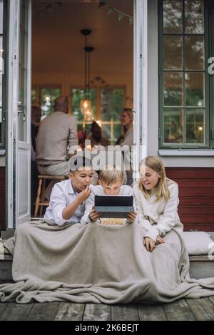 Aufgeregt Freunde mit Decke Film auf digitalem Tablet zu sehen, während sie auf der Veranda sitzen Stockfoto