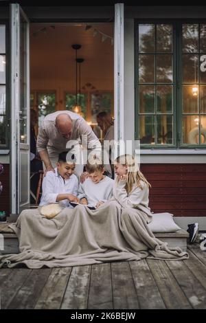 Mann, der mit Kindern auf einem digitalen PC spricht, während er auf der Veranda sitzt Stockfoto