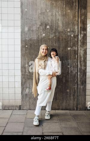 Porträt einer glücklichen Frau, die ihre Tochter trägt, während sie vor der Wand steht Stockfoto