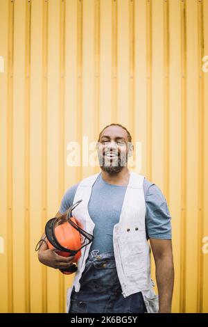 Porträt eines glücklichen, reifen Bauarbeiters, der mit einem Hardhut gegen eine gelbe Metallwand steht Stockfoto