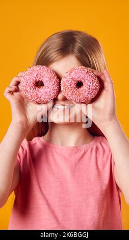 Studio Portrait Of Girl Halten Zwei Donuts Vor Den Augen Gegen Gelben Hintergrund Stockfoto