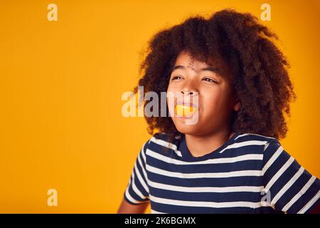 Studio Portrait Of Smiling Boy Pulling Funny Face With Orange For Mouth Against Yellow Background Stockfoto