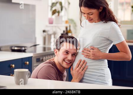 Aufgeregt schwanger Transgender Paar zu Hause in der Küche mit Mann hören Baby Herzschlag Stockfoto