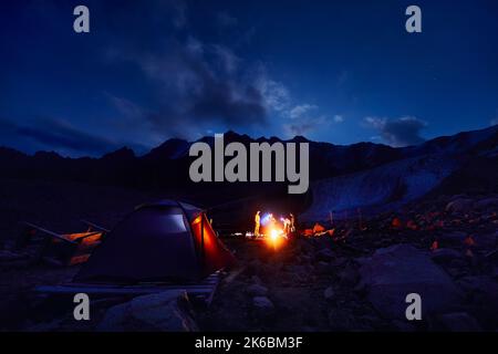 Touristen mit Scheinwerfern in der Nähe von beleuchteten Zeltlagern in der Nähe von Lagerfeuer in den Schneebergen unter Nachthimmel mit Sternen. Stockfoto