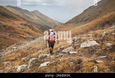 Portrait eines alten bärtigen Wanderers mit großem Rucksack und Wanderstöcken, der gegen das Bergtal läuft. Outdoor- und Trekkingkonzept. Stockfoto
