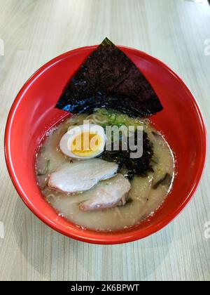 Aus nächster Nähe sehen Sie heiße Hühnchen-Ramen-Nudelsuppe mit aufschneidendem Ei, Hähnchen, Gemüse und Seetang in roter Schüssel. Traditionelle japanische Küche. Stockfoto