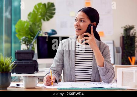 Weibliche Architektin, Die Im Büro Am Schreibtisch Sitzt Und Auf Dem Mobiltelefon Spricht Stockfoto