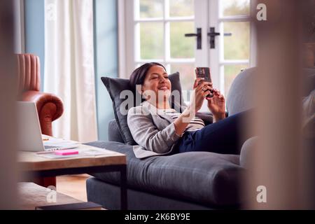 Frau, die von zu Hause aus eine Pause nimmt und auf dem Sofa liegt, um das Mobiltelefon zu überprüfen Stockfoto