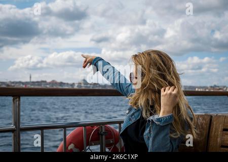 Blonde langhaarige Frau, die mit der Fähre in Bosporus reist und mit der Hand auf einen Punkt zeigt. Stockfoto