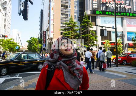 Portait einer jungen muslimischen Frau, die mit dem Yodobashi-Akiba-Gebäude lächelt, und Menschenmenge, die in Akihabara spazieren und einen klaren blauen Himmelshintergrund haben. Stockfoto