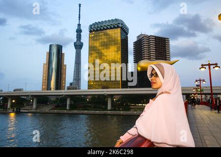 Porträt einer muslimischen Frau, die auf der Azuma-Brücke steht, mit übersinnlichem Gesichtsausdruck und dem Gebäude der Asahi-Gruppe und dem Sumida-Fluss bei Sonnenuntergang. Stockfoto