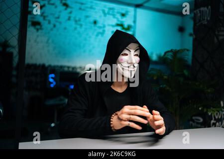 Nahaufnahme eines gefährlichen Hackers, der eine anonyme Maske und einen Kapuzenpullover trägt und Perlen in den Händen hält und an den Tisch im Kellerraum blickt. Stockfoto