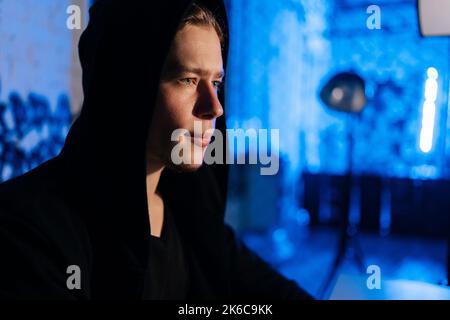 Nahaufnahme eines gutaussehenden gesuchten Hackers in einem Sweatshirt mit Kapuze, der in Sicherheitssysteme hackt und in einem dunklen Raum mit blauem Neon sitzt Stockfoto
