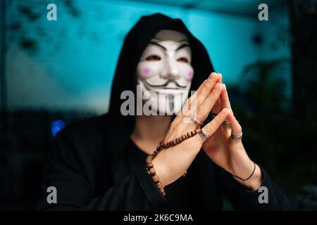 Nahaufnahme eines gefährlichen Hackers, der eine anonyme Maske und einen Kapuzenpullover trägt und Perlen in den Händen hält und an den Tisch im Kellerraum blickt. Stockfoto