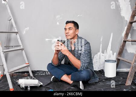 Ein attraktiver junger asiatischer Mann sitzt auf dem Boden und macht eine Kaffeepause vom Heimwerker Stockfoto