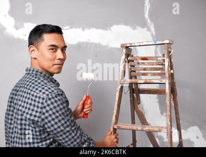Ein junger asiatischer Mann sieht ernst aus, als er sich darauf vorbereitet, rissige Wände zu malen Stockfoto