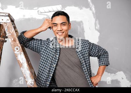 Ein attraktiver asiatischer Mann lächelt und posiert mit einem Farbroller gegen die Wand Stockfoto