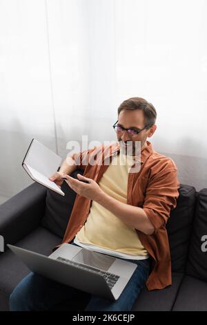 Lächelnder Mann in einer Brille, der während eines Videoanrufs auf dem Laptop zu Hause auf ein leeres Notebook zeigt, Stockbild Stockfoto