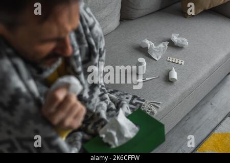 Kranker Mann mit Papierservietten sitzt auf der Couch in der Nähe von elektronischen Thermometer und Behälter mit Behandlung, Stock Bild Stockfoto