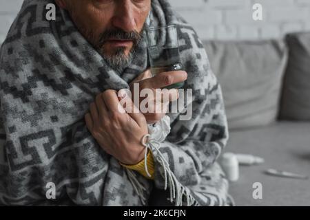 Teilansicht des Mannes, der in eine warme Decke gehüllt ist und ein Glas Wasser hält, Stockbild Stockfoto