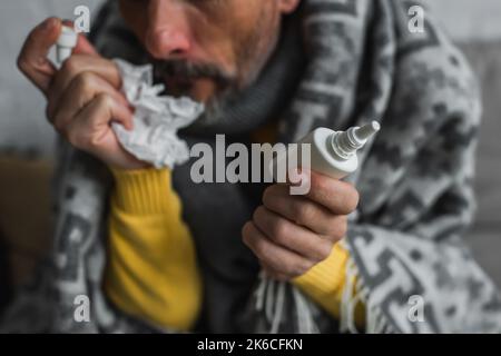 Teilansicht eines verschwommenen Mannes, der Nasenspray und zerknitterte Papierservietten hält, während er an laufender Nase leidet, Stockbild Stockfoto