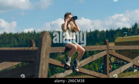 Junge Fotografin schießt Landschaft mit Digitalkamera auf schönen grünen Hügel, sitzt auf Holzzaun, fotografieren die Natur. Mädchen füllen das Portfolio mit erstaunlichen Fotos. Stockfoto