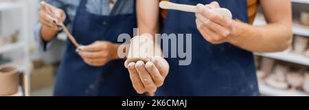 Beschnittene Ansicht des Handwerkers, der in der Nähe einer verschwommenen afroamerikanischen Frau in der Werkstatt einen Shaper und ein Tonprodukt hält, Banner, Stockbild Stockfoto