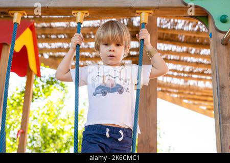 Ein Kind klettert an einem heißen Sommertag in einem Park auf einem Spielplatz auf ein alpines Netz. Kinderspielplatz in einem öffentlichen Park, Unterhaltung und Erholung f Stockfoto