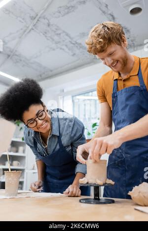 Lächelnder Mann, der in der Nähe afroamerikanischer Kunsthandwerkerin im Töpferstudio einen Keramikbecher macht, Stockbild Stockfoto