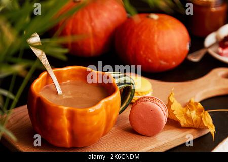 Herbsttasse aus Kürbis Latte in Form von Kürbis, Makronen zwischen Kürbis und Blättern. Stimmungsvolle, gemütliche Kaffeezeit. Stockfoto