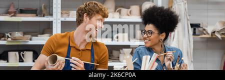 Lächelnder Rotschopf Mann Malerei auf Ton Produkt in der Nähe afroamerikanische Freundin in Töpferei, Banner, Stock Bild Stockfoto