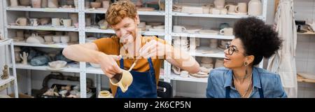 Rotschopf Mann Malerei auf Tonbecher in der Nähe fröhliche afroamerikanische Freundin in Töpferwerkstatt, Banner, Stock Bild Stockfoto