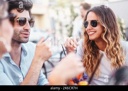 Lächelnde Erwachsene Männer und Frauen in Sonnenbrillen, die Spaß haben, am Pub-Tisch zu sitzen und sich zur Happy Hour zu unterhalten - trendige, fröhliche Freunde, die sich bei s zusammenschließen Stockfoto