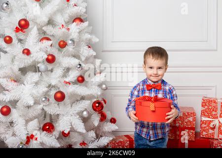 Kleiner niedlicher Junge in lässiger Kleidung, der in einem weißen Raum zwischen weihnachtsdekorationen steht. Weihnachtsfoto des Kindes im dekorierten Weihnachtszimmer. Neujahr Konz Stockfoto