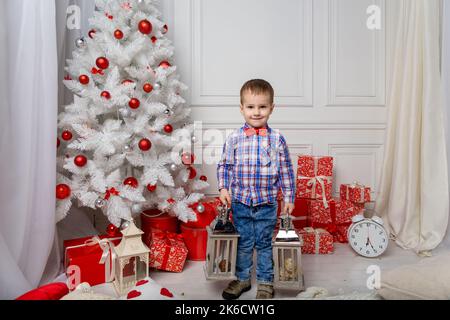 Kleiner niedlicher Junge in lässiger Kleidung, der in einem weißen Raum zwischen weihnachtsdekorationen steht. Weihnachtsfoto des Kindes im dekorierten Weihnachtszimmer. Neujahr Konz Stockfoto