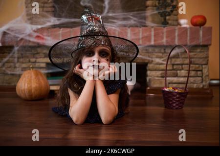 Frohe Halloween! Bezauberndes mystisches Mädchen, in einer Hexe gekleidet, liegt auf dem Boden vor einem mit Spinnennetz bedeckten Kamin Stockfoto