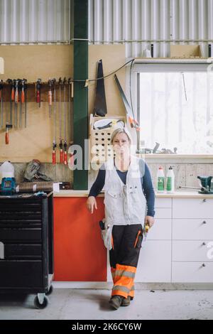 In voller Länge lächelnde, reife Tischlerin, die in der Werkstatt an der Werkbank steht Stockfoto