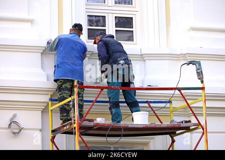 Arbeiter reparieren die Gebäudewand, die auf einer Hebebühne steht. Zwei Bauherren lackieren die Hausfassade, Bau- und Reparaturarbeiten Stockfoto