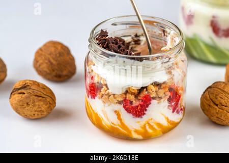 Glas und Teil der Sekunde mit Joghurt, Himbeere, Müsli, Mango- und Pistazien-Creme, ganze Walnuss auf hellem Hintergrund, Nahaufnahme. Stockfoto