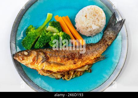 Gebratene Forelle gefüllt mit Zwiebeln, Gemüsegarnitur (Brokkoli und Karotte) und Reis auf blauem Teller, Nahaufnahme. Stockfoto