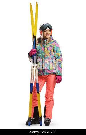 Ganzkörperporträt einer jungen Frau mit einem Helm, der steht und ein Paar Ski auf weißem Hintergrund hält Stockfoto