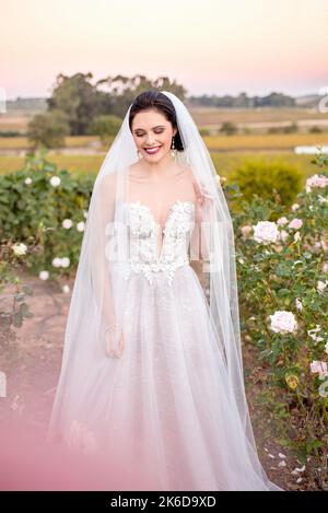 Wunderschöne dunkelhaarige Braut in einem Blumenhochkleid mit Spitze und einem langen Schleier, der in einem weißen Rosengarten lächelt. Stockfoto