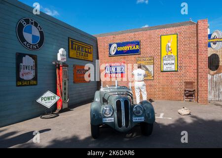 Das BMW-Auto Frazer Nash parkte neben einer Wand mit authentischen Auto-Werbeschildern beim BARC Revival Meeting, Goodwood Motorrennstrecke, Chichester, Großbritannien Stockfoto