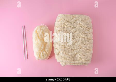 Flaches Lay mit Garnknäuel, Nadeln und Strickschal auf hellrosa Hintergrund. Handgefertigtes und stricktes Hobby-Konzept Stockfoto