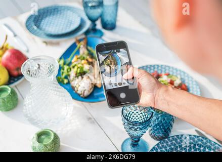 Frau macht Foto mit Smartphone gegrillten Fisch im Ofen und serviert auf einem weißen Holztisch mit Gemüsesalat und Obst. Gesunde Zubereitung von Meeresmahlzeiten Stockfoto