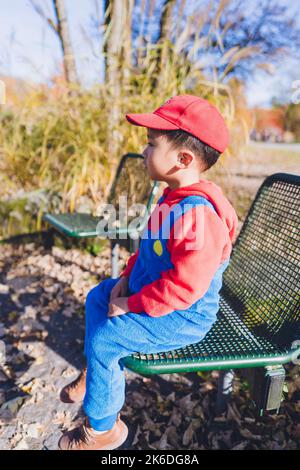 Ein in Kostüm gekleideter Junge sitzt auf einer Bank in einem öffentlichen Park. Stockfoto