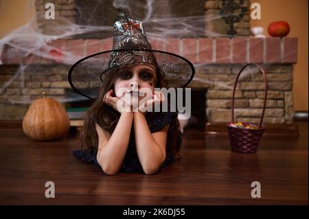 Frohe Halloween. Bezauberndes mystisches Mädchen, in einer Hexe gekleidet, liegt auf dem Boden vor einem mit Spinnennetz bedeckten Kamin Stockfoto