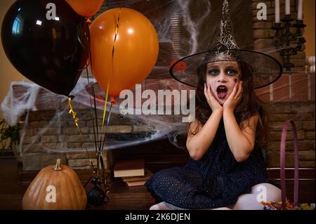 Charmantes kleines Mädchen, in Hexe gekleidet, sitzt in der Nähe von orangefarbenen Ballons und Kürbis vor einem mit Spinnennetz bedeckten Kamin Stockfoto